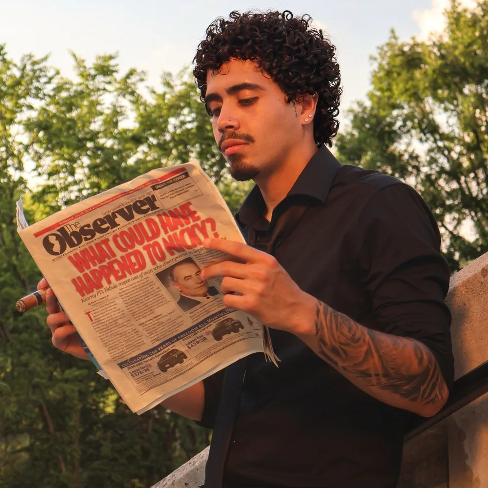 Dru reading a newspaper, a black-and-grey sleeve on his forearm